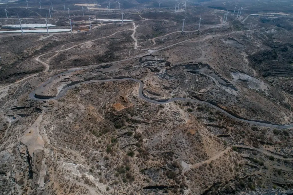 Aerial shot of a winding road amidst arid, rocky landscapes with wind turbines in the background.